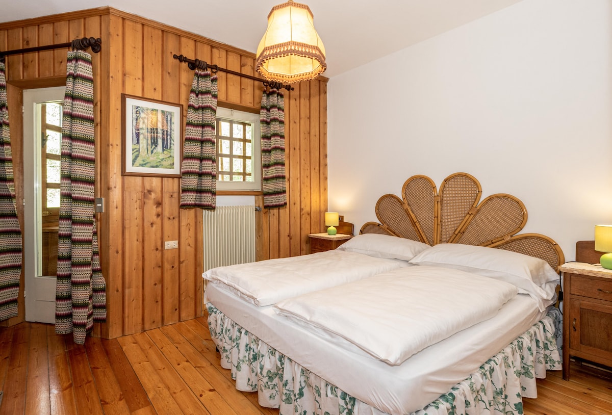 A cozy bedroom features a spacious bed with a decorative headboard, positioned against wooden paneling. Natural light enters through a window adorned with patterned curtains. Nightstands with lamps provide soft illumination, enhancing the room's inviting atmosphere.