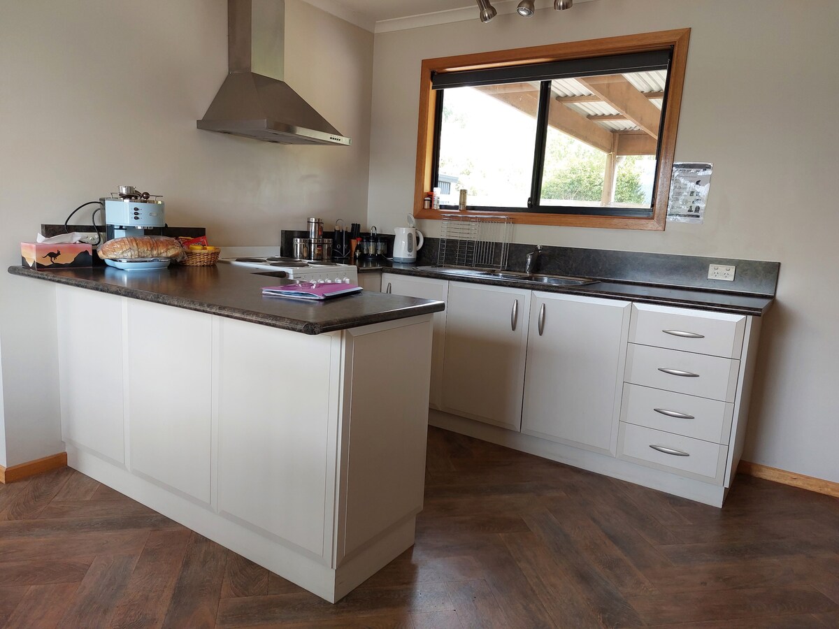 A functional kitchen space is highlighted by a central island with a dark countertop. Stainless steel appliances are positioned alongside ample cabinetry. Large windows provide natural light, offering views of the outdoor area. Cooking utensils and a coffee maker are neatly arranged on the counter.