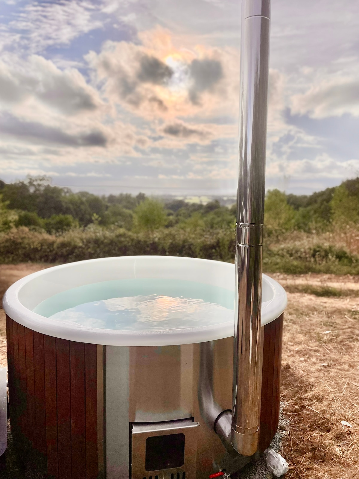 A wooden hot tub is situated on an elevated area, featuring a shiny metallic chimney. The calm water surface reflects the soft clouds and sunlight above, providing a view of the surrounding greenery and distant hills.