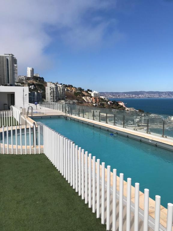A rooftop pool extends along a barrier, surrounded by lush green grass. The water sparkles under sunlight, offering panoramic views of the coastline and city. Tall buildings line the hills in the background, and a clear blue sky complements the serene setting.