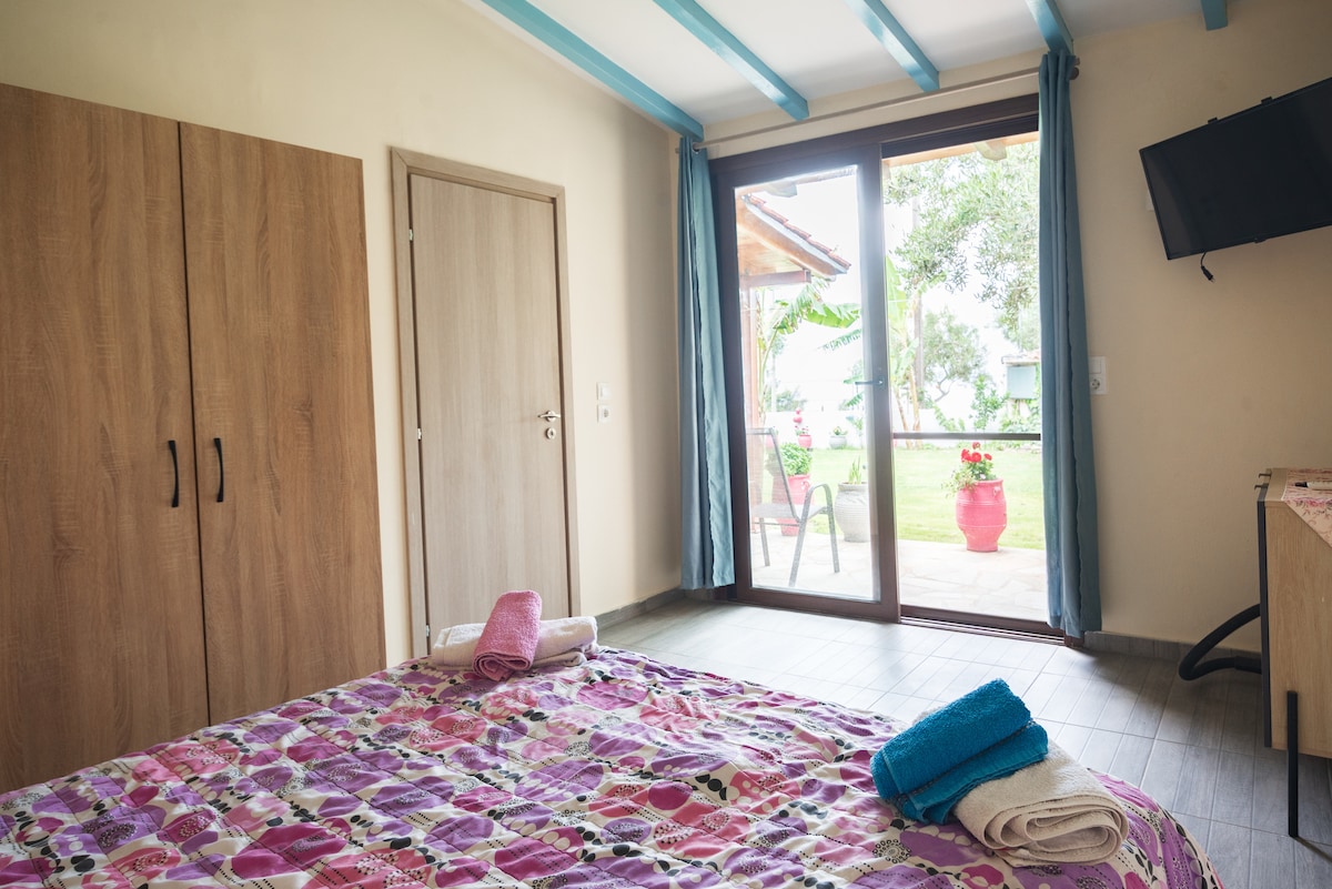 A spacious room features a large bed covered with a colorful patterned blanket. Two towels are neatly placed on the bed. A glass door opens to a private outdoor area with vibrant plants, while a wall-mounted TV is visible in the corner.