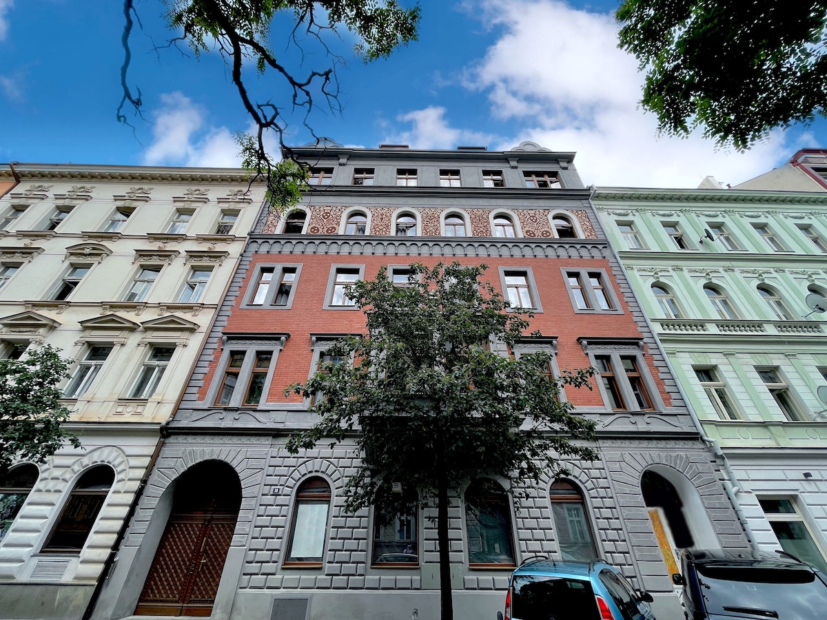 A historic building is showcased with a blend of brick and stone finishes, featuring an ornate facade. Tall windows and a variety of architectural details highlight the elegant structure. Nearby trees provide a touch of greenery against the clear blue sky.