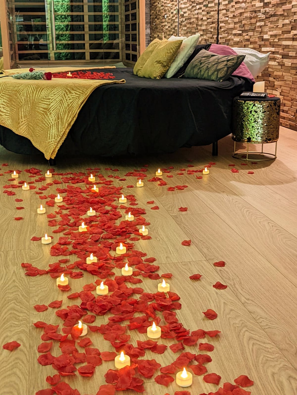 A spacious round bed is surrounded by decorative pillows, adding color to the dark bedding. A pathway of red rose petals leads towards the bed, with flickering candles illuminating the floor, creating a serene and romantic setting within a warm wooden interior.