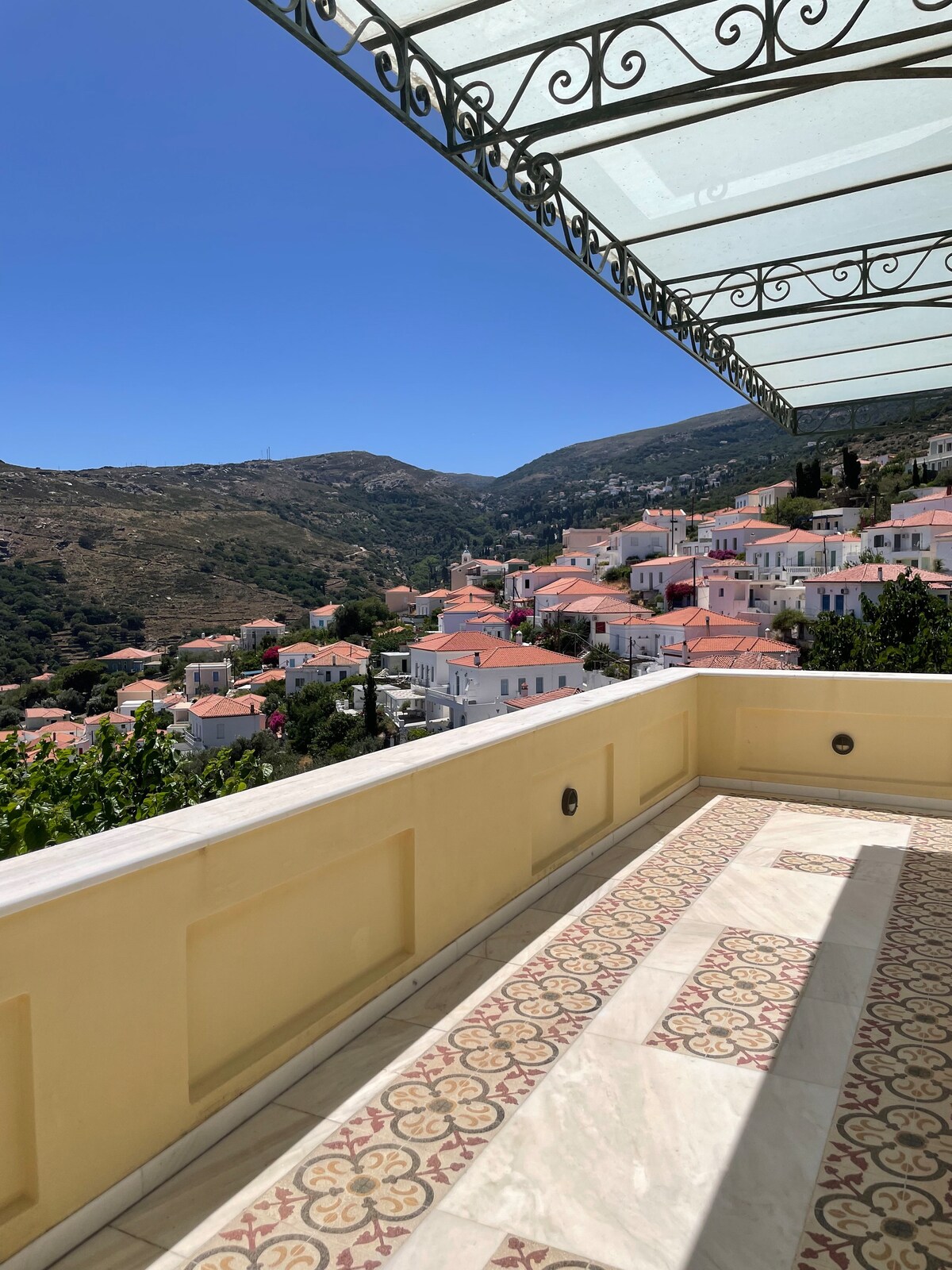 A spacious balcony showcases intricate tiled flooring and a covered area with a wrought-iron canopy. Stunning views of the surrounding hills and village with white and pastel-colored buildings are visible, set against a clear blue sky.