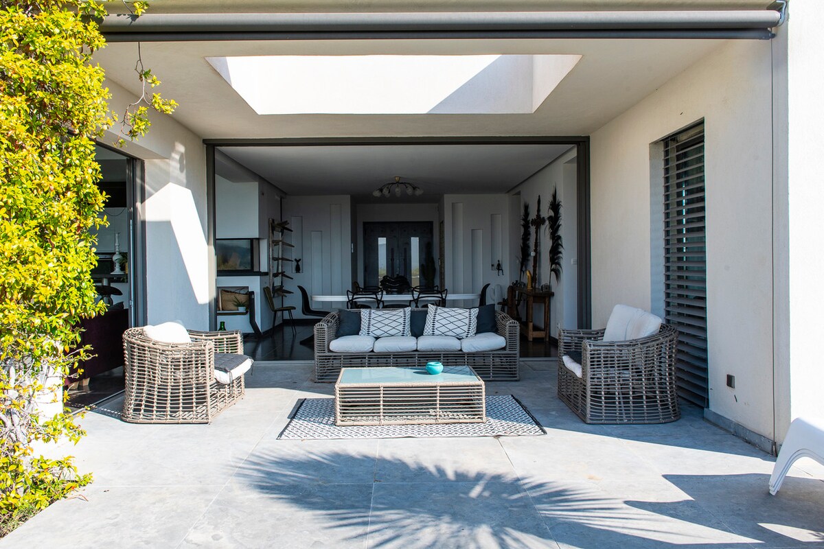 An outdoor lounge area features comfortable seating arranged around a central coffee table. The space is framed by large glass doors, allowing for a seamless transition between indoor and outdoor living. A serene courtyard with greenery is visible in the background.