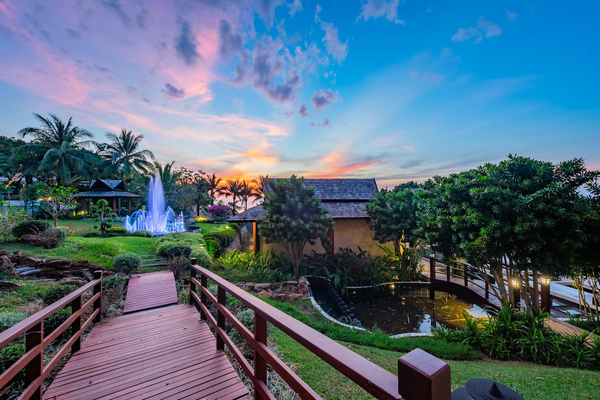 A wooden pathway leads towards a tranquil garden setting at sunset, where a gentle fountain is illuminated in soft colors. Lush greenery surrounds a serene koi pond, with a detached villa visible in the background under a colorful sky.