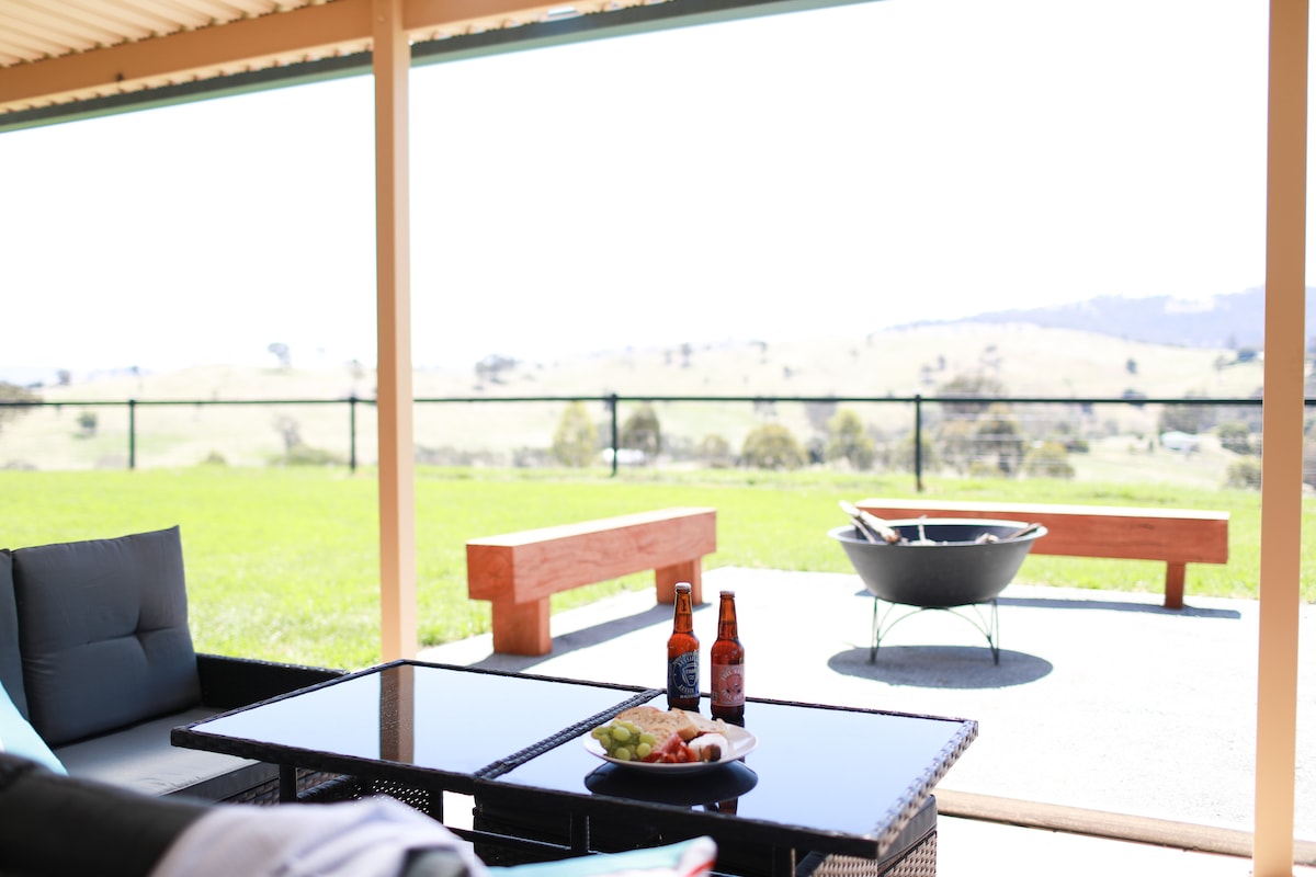 An outdoor seating area features a low table with a plate of snacks and two bottles of drink. Beyond the space, a fire pit is visible, surrounded by manicured grass and offering views of rolling hills in the distance.