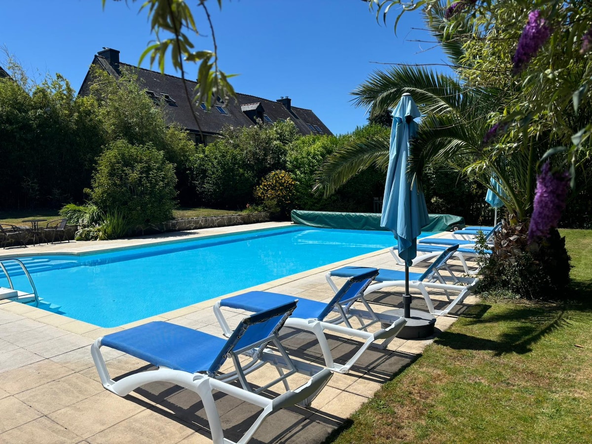 A heated outdoor pool is surrounded by lush greenery, featuring several sun loungers arranged on a paved area. An umbrella provides shade next to the pool. The clear blue water reflects the sunny sky, creating a serene environment for relaxation.