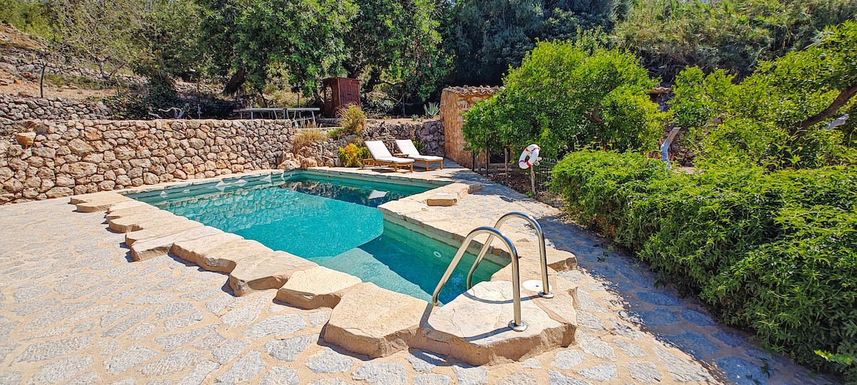 A natural stone pool is surrounded by lush greenery and decorative rocks. Sun loungers are positioned neatly beside the water, enhancing the outdoor space. Mature trees provide shade, while a stone wall offers privacy, creating a serene atmosphere for relaxation.