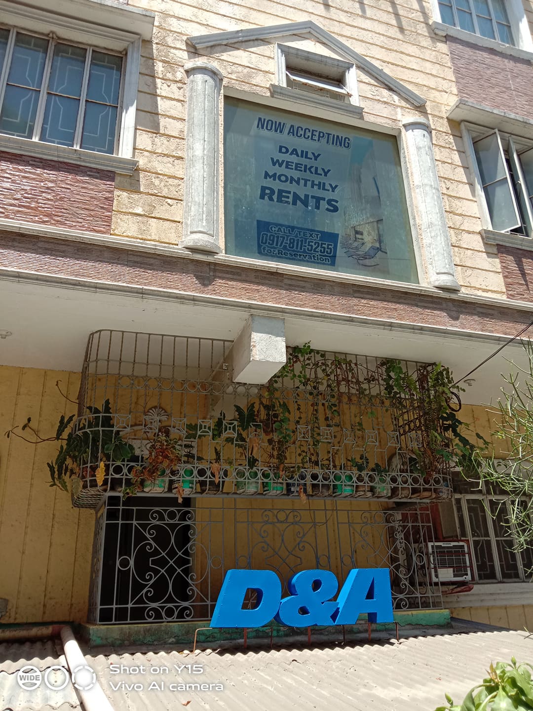 The exterior of a five-story building is showcased, featuring yellow and brown walls. A large banner indicates availability for daily, weekly, and monthly rentals. A metal gate adorned with plants is visible in the foreground, complemented by a prominent blue sign with the letters 'D&A.'