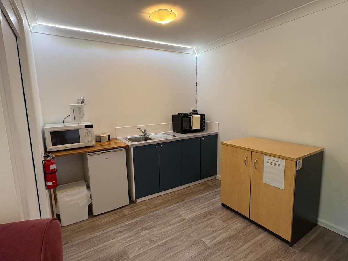 A compact kitchenette is displayed with a sink, microwave, and refrigerator. Storage cabinets in a muted tone are also present. A fire extinguisher is mounted on the wall, ensuring safety. Natural light illuminates the space from overhead lighting.