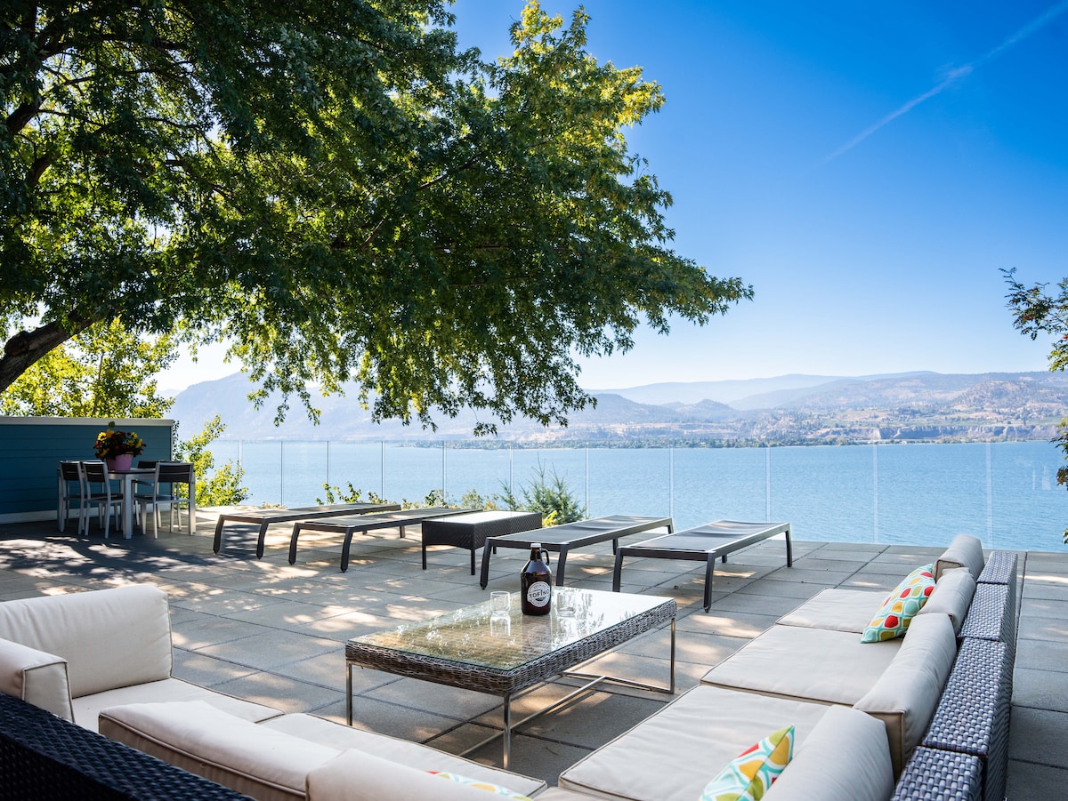 An expansive deck overlooks the water, featuring multiple lounge chairs and a sectional sofa with colorful cushions. A glass table sits central on the deck, complementing the views of the lake and surrounding hills under clear blue skies.