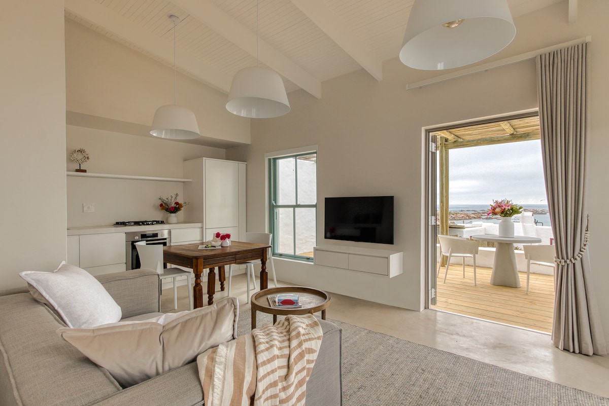 A bright and airy living area features a spacious gray sofa adorned with a cozy throw, positioned near a wooden dining table. A flat-screen television is mounted on the wall, while large windows open to a patio area, providing views of the outdoor dining setup and sea beyond.