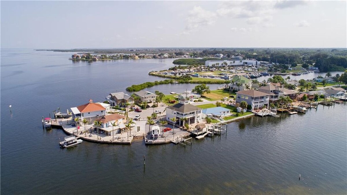 An aerial view captures the serene waterfront community, featuring several homes along the shoreline. Calm waters reflect the landscape, while boats are docked at various piers. Lush greenery lines the edges, creating a peaceful and inviting atmosphere.