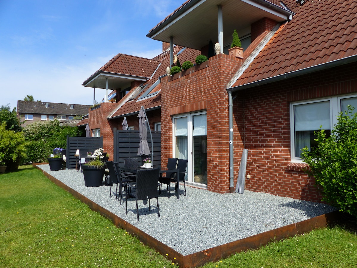 An outdoor terrace is presented, featuring a gravel surface and dark patio furniture. Potted plants add greenery alongside the wall. The building's brick exterior and multiple windows are visible, creating a welcoming outdoor space. Sunshade umbrellas provide comfort for guests.