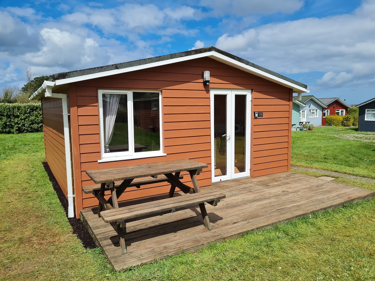 A wooden cabin with a warm orange exterior is presented. A wooden picnic table is situated on the deck in front of large windows, allowing natural light to illuminate the interior. Lush green grass surrounds the cabin, contributing to a welcoming outdoor setting.