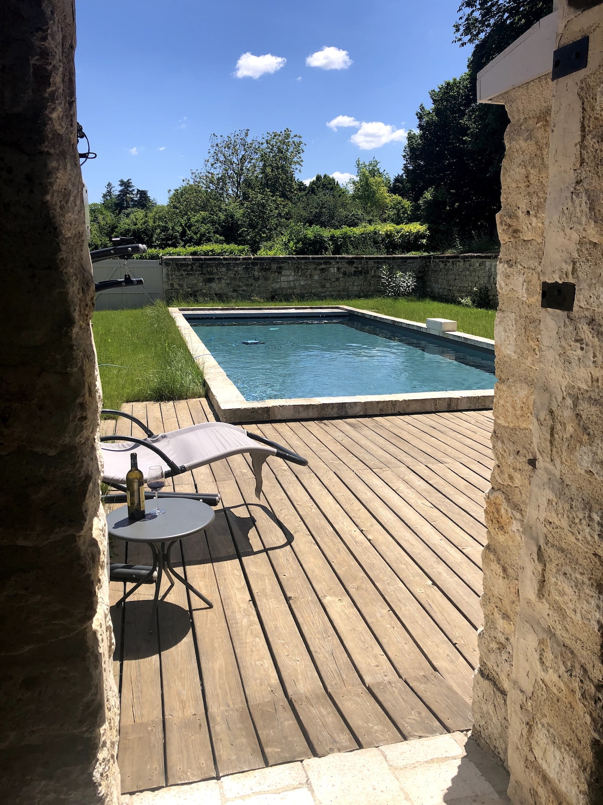 A view of the outdoor area from the entrance, showcasing a swimming pool surrounded by wooden decking. Lounge chairs are positioned nearby, and lush greenery borders the space, enhancing the serene environment under a clear blue sky.