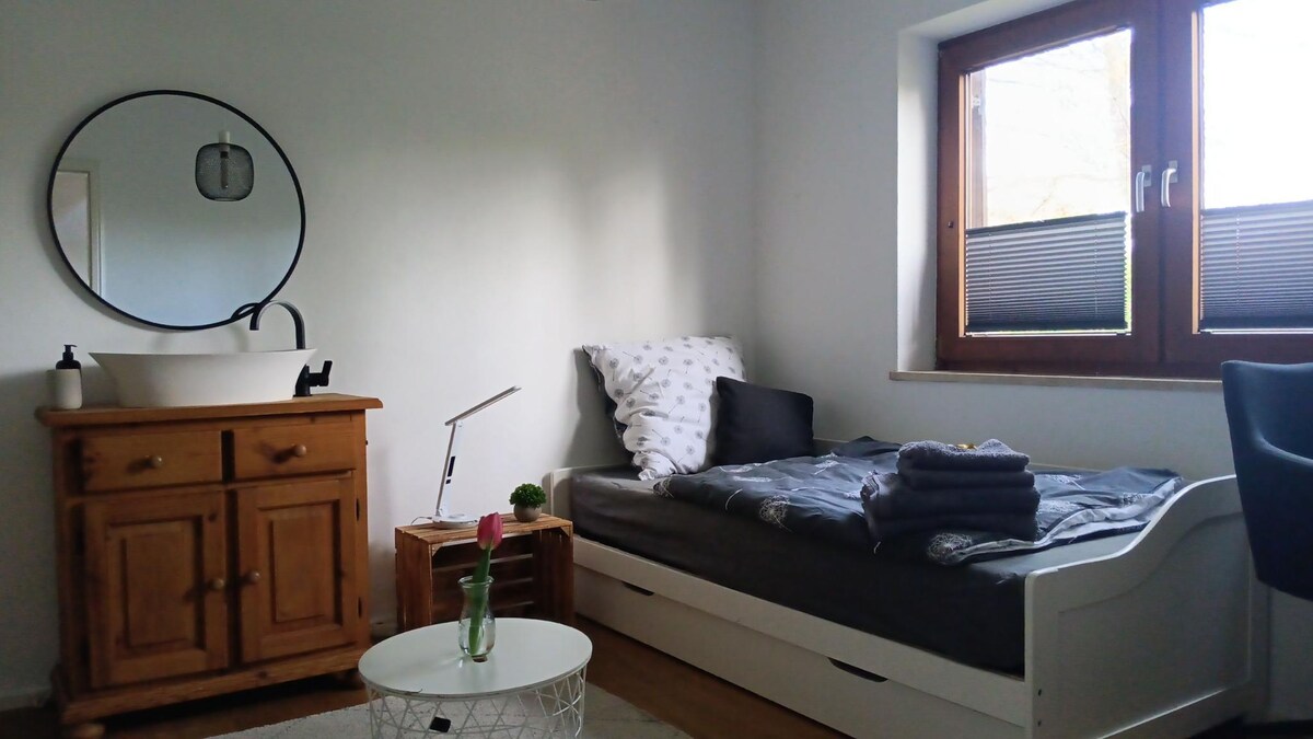 A light-filled room features a single bed with gray linens and neatly stacked towels. A round mirror hangs above a wooden cabinet with a sink. A small coffee table sits in front, accompanied by a chair. The window offers views of the natural surroundings.