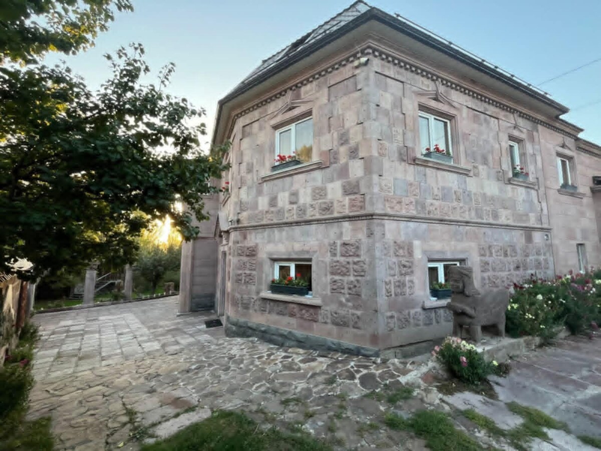 The exterior of a stone guest house is showcased, featuring a robust design with a lightly textured facade. Flower boxes adorn several windows, complemented by a surrounding garden. A patterned stone pathway leads up to the entrance, framed by greenery and blooming flowers.