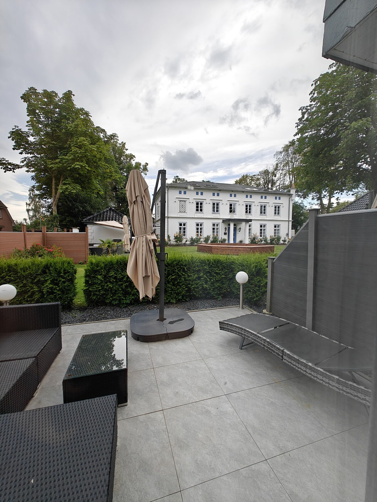 A private outdoor seating area features a lounging chair and a small table under a large umbrella. The view reveals a well-maintained garden and a restored historic building in the background, surrounded by greenery. Soft lighting elements are visible, enhancing the relaxed setting.