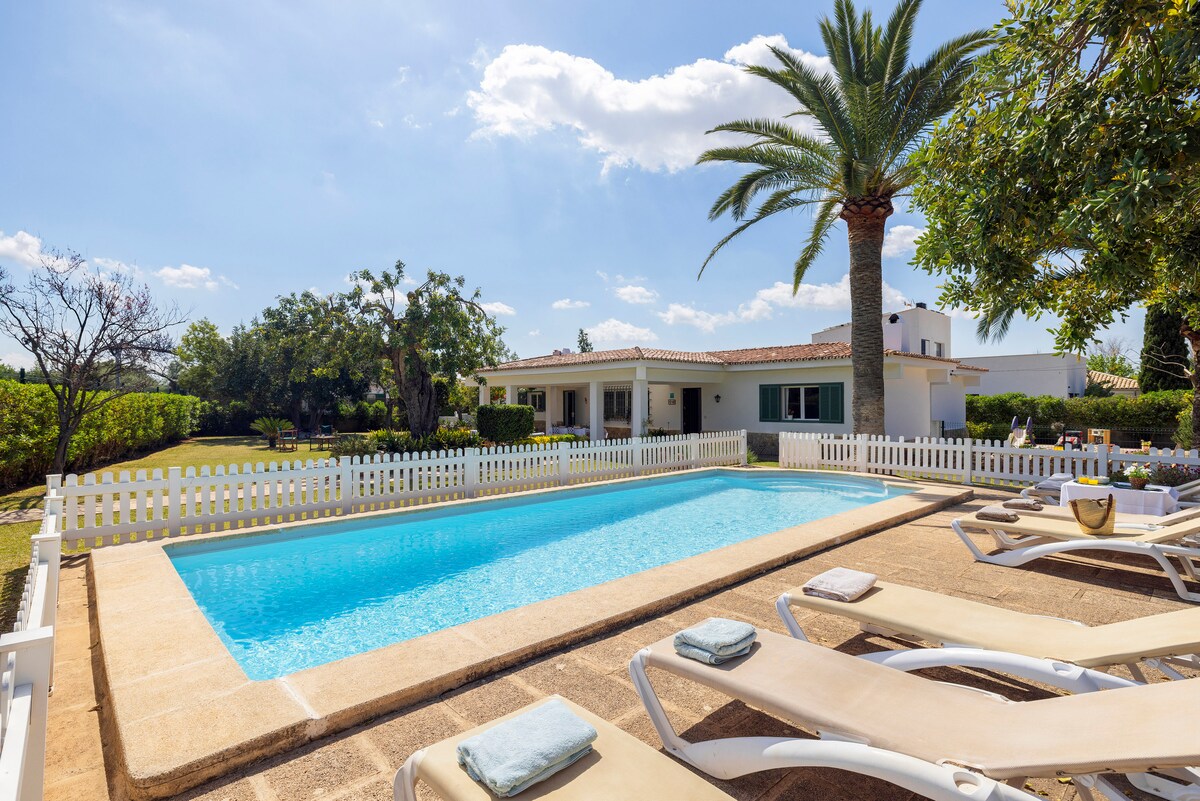 A serene outdoor area features a clear swimming pool surrounded by sun loungers. A well-maintained garden and palm trees provide shade, while a white fence offers a boundary. The spacious house is visible in the background beneath a bright blue sky.