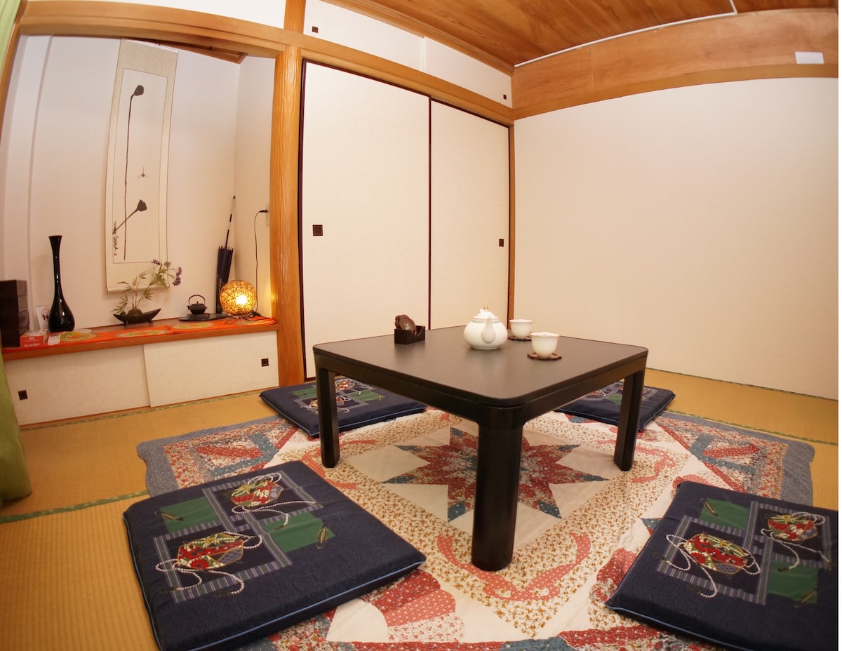 An elegantly arranged traditional room features a low, dark wooden table surrounded by floor cushions. A decorative alcove displays a beautiful vase and art piece. Tatami mats cover the floor, while sliding doors create a serene ambiance in this inviting space.
