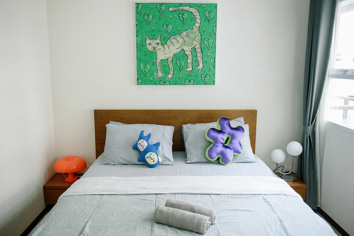 A cozy bedroom features a large bed dressed in light grey linens. Colorful decorative pillows rest against the wooden headboard. Above the bed, a vibrant cat painting adds a playful touch. Soft lighting is provided by a bedside lamp and a small light fixture.