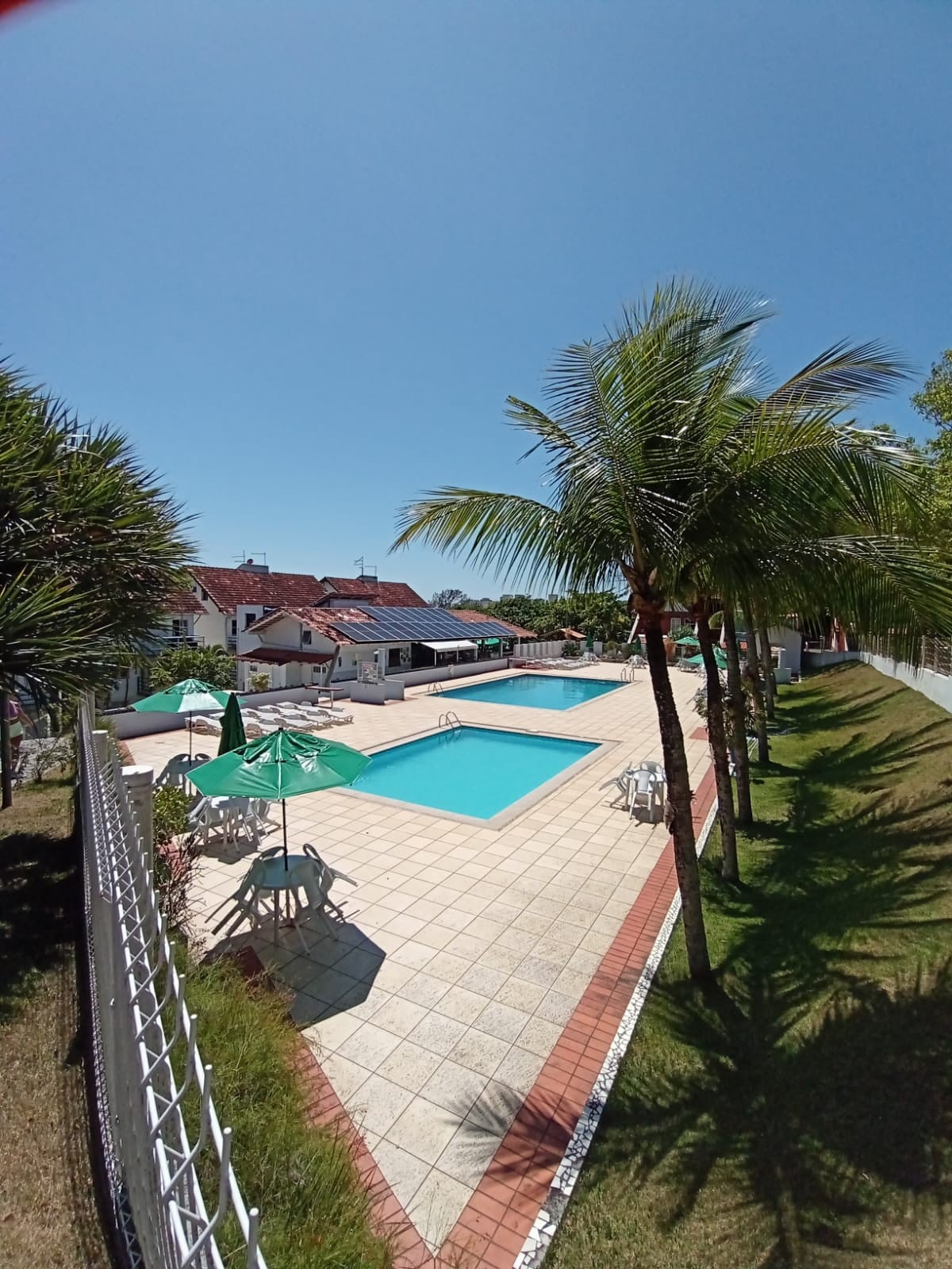 A clear view of a bright outdoor pool area is presented, featuring two swimming pools surrounded by well-maintained landscaping. Lounge chairs and umbrellas provide shaded seating options. Palm trees contribute to a relaxed ambiance in the sun-drenched space.