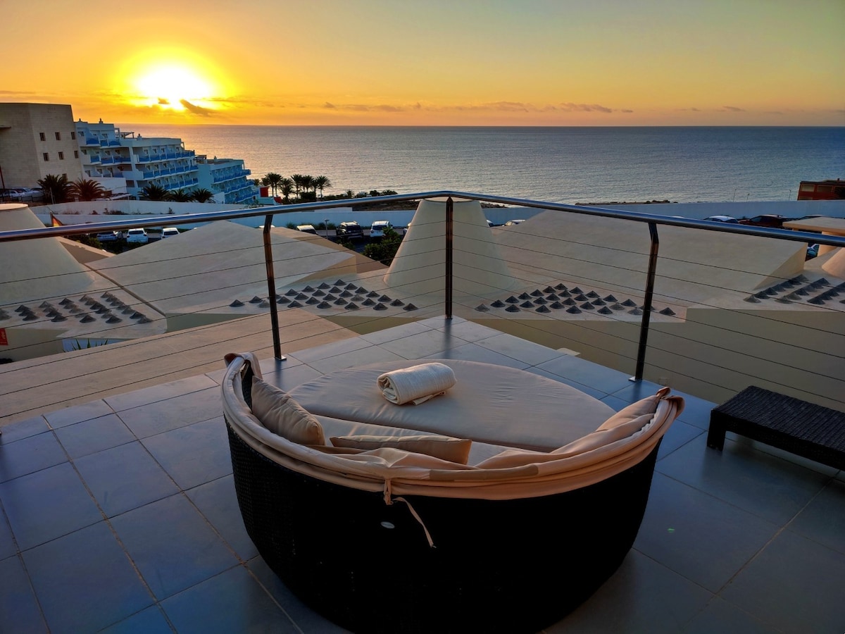 A private terrace offers a sweeping view of the ocean at sunset. A circular lounge bed is positioned on the terrace, accompanied by a rolled towel. The horizon features soft hues of orange and pink, blending with the calm sea.