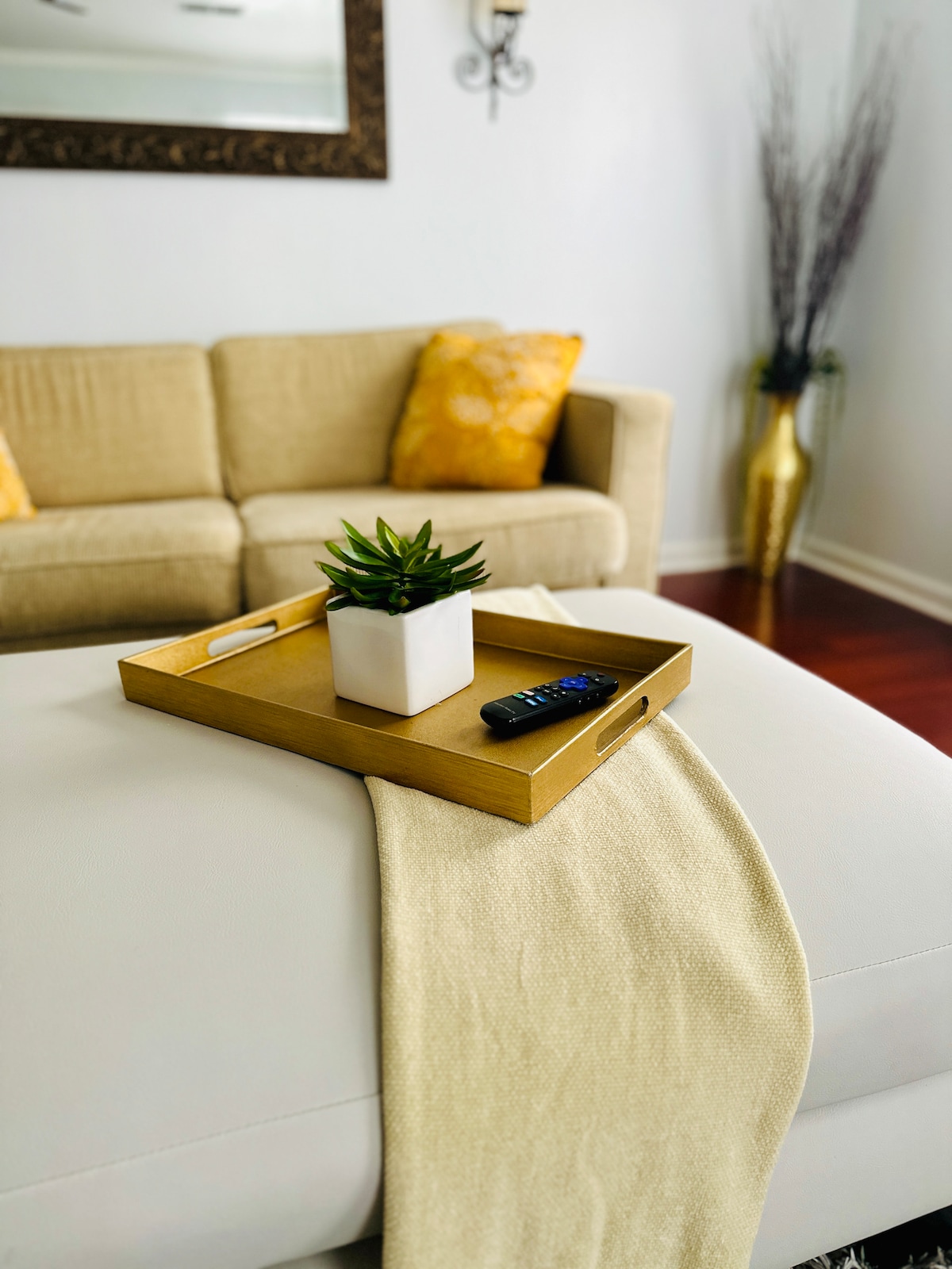 A light-colored ottoman features a wooden tray holding a potted plant, accompanied by a remote control. Soft golden accents are present on the nearby couch, which is positioned against a neutral-toned wall, while a decorative vase adds elegance to the scene.