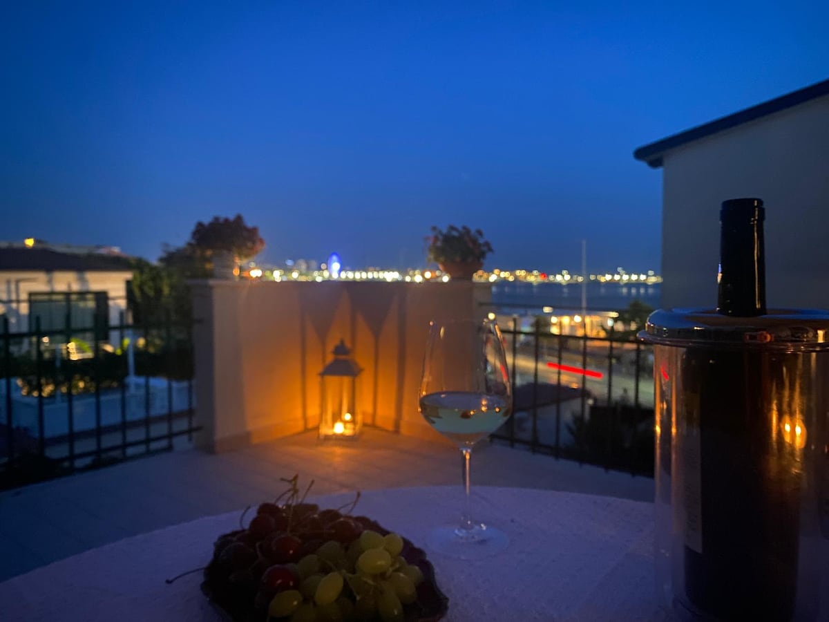 A terrace is shown at night, featuring a table set with a glass of white wine and a plate of mixed grapes. Soft lighting is provided by a nearby lantern, while the city skyline sparkles in the distance, creating a serene ambiance.