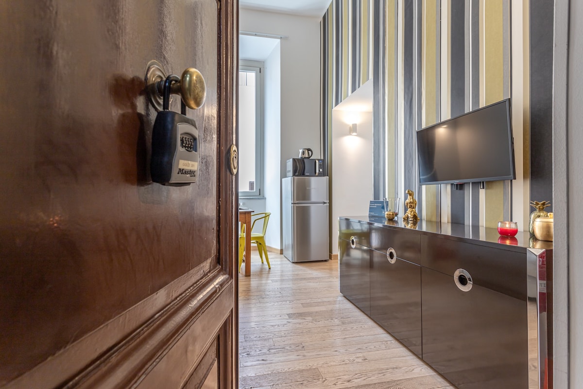 A view from the entrance showcases a modern living space featuring striped wallpaper and light wooden flooring. A sleek TV stand stands with decorative items, while a compact kitchen area is visible in the background alongside a refrigerator. A dining table and chairs add to the functionality of the room.