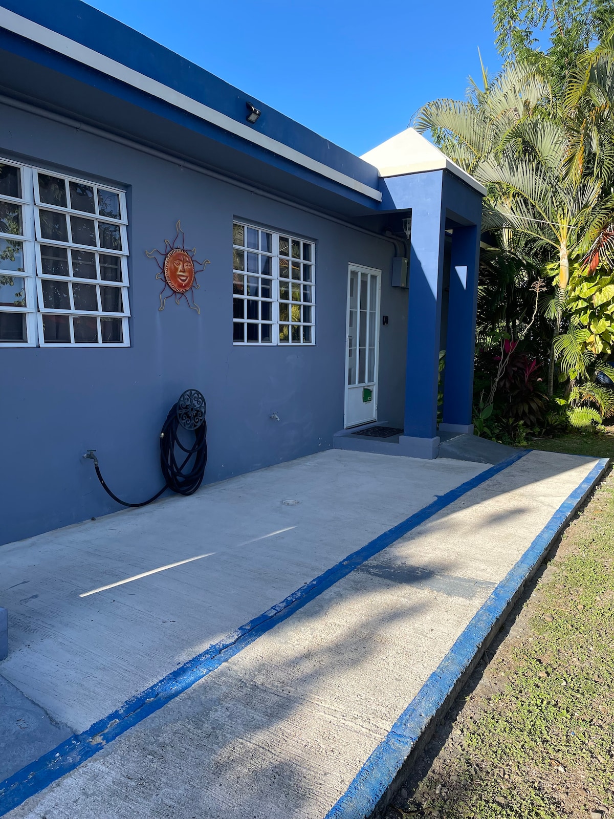 The exterior of a blue home is displayed, featuring a concrete pathway leading to a white door. Multiple windows allow natural light, and a sun motif is visible on the wall. Lush greenery surrounds the area, adding to the inviting atmosphere.