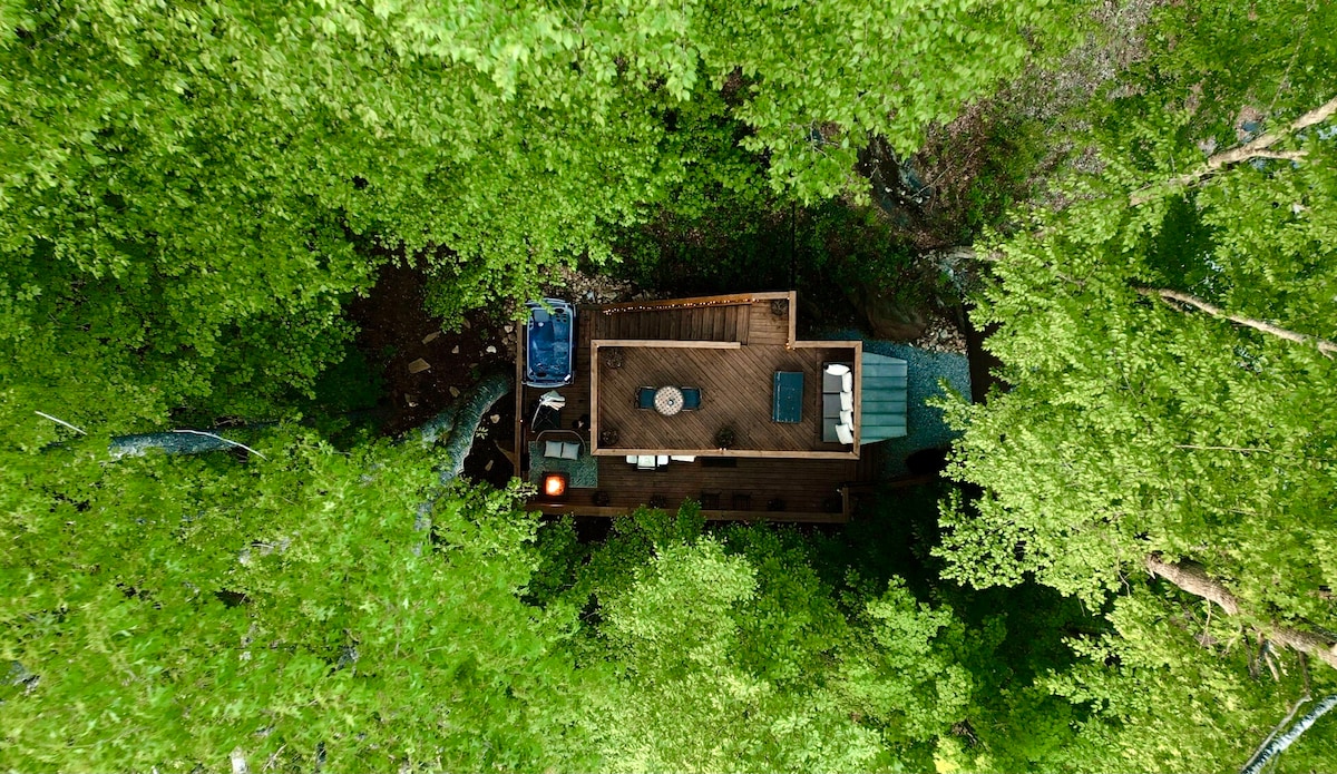 An aerial view of the Tiny Bunker shows the structure nestled within lush greenery. The outdoor deck features a hot tub, dining area, and lounge furniture, all surrounded by trees, creating a sense of seclusion and a connection with nature.