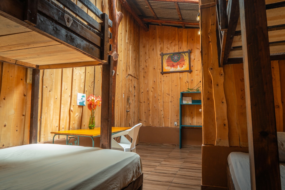 A spacious room features wooden walls and high ceilings, creating a warm atmosphere. A lower bunk bed is present alongside a double bed. A wooden table with fresh flowers and a blue shelf add functionality, while a colorful artwork decorates the wall.