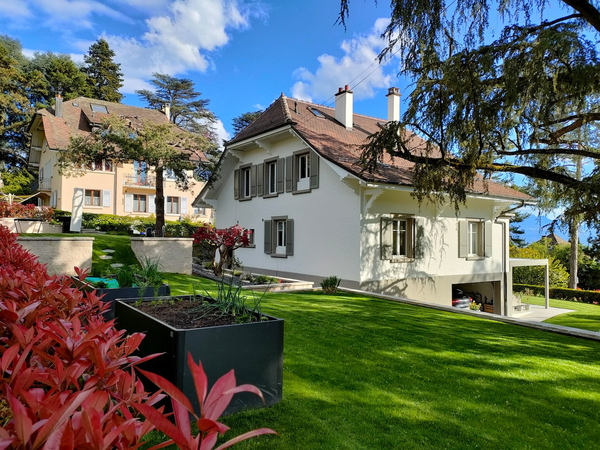 A charming white house is surrounded by well-maintained green lawns and vibrant red foliage. The structure features classic windows and a sloped roof, complemented by a landscaped garden. The background reveals additional properties and a clear blue sky.