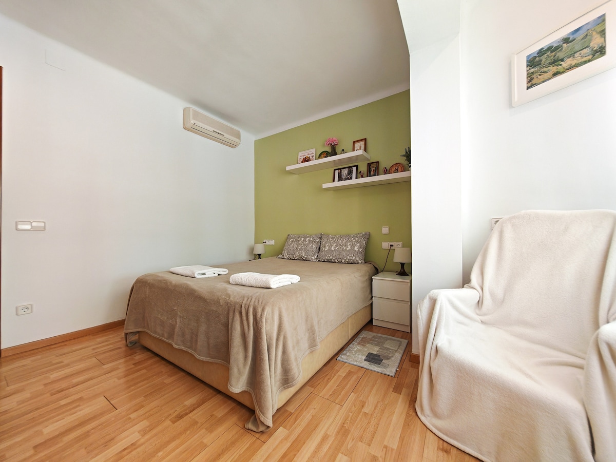 The bedroom features a spacious bed with light-colored bedding and decorative pillows. A soft throw is draped over the side of the bed. Shelves above the bed display small plants and personal items. A cozy chair is positioned in the corner, enhancing the inviting atmosphere.