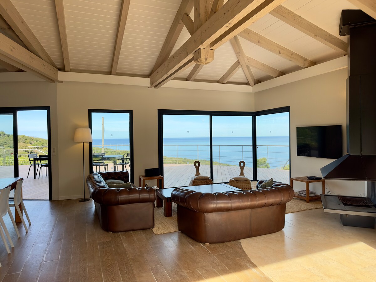 A spacious main living area is highlighted by large glass doors, providing unobstructed views of the sea. The room features a leather sofa arrangement and a modern fireplace. Natural light fills the space, complemented by a warm wood ceiling and minimalist decor.