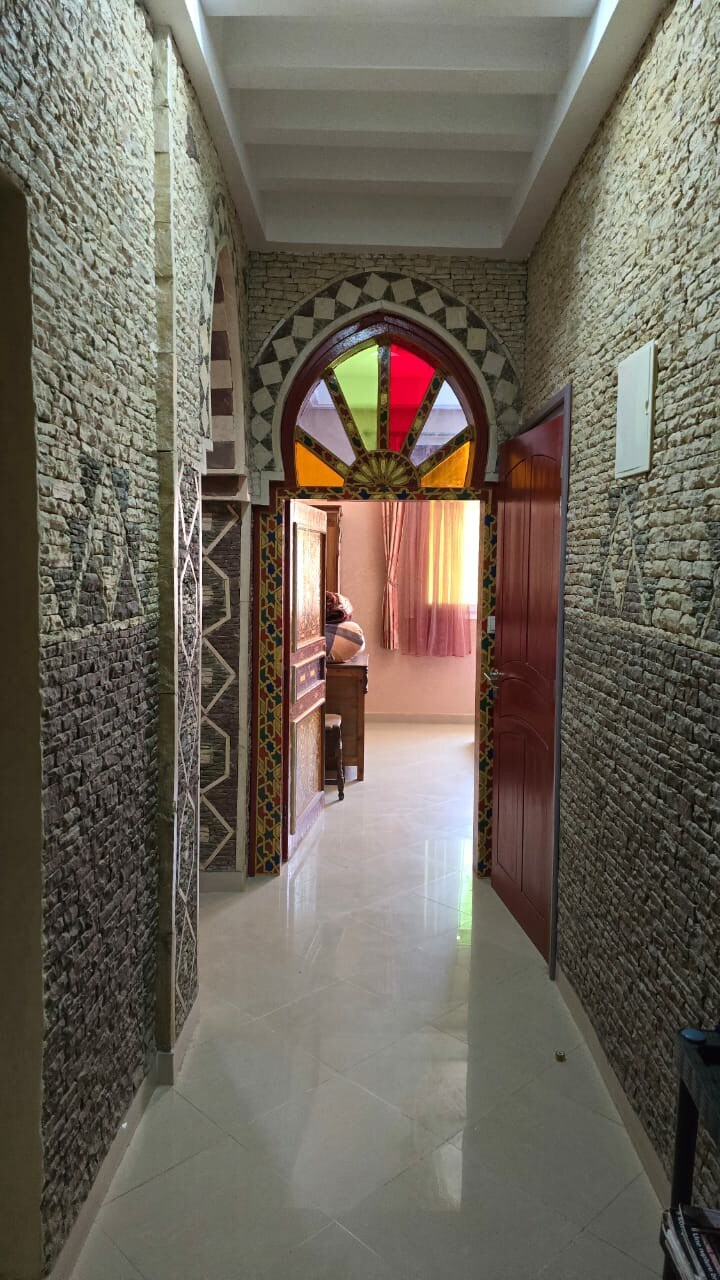 A hallway showcases intricately patterned stone walls, framed by bright colored window panes. Two doors can be seen leading to private spaces, while soft natural light illuminates the room, highlighting the decorative elements along the corridor.