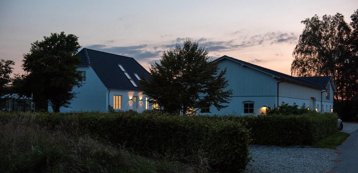 The exterior of a two-story building is visible, featuring large windows that softly illuminate the space. A gently sloping garden with hedges surrounds the property, set against a dusky sky with hints of sunset. A peaceful rural atmosphere is suggested by the nearby trees.