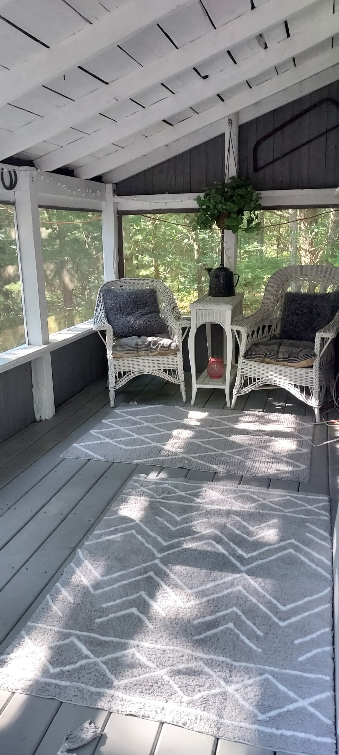 A screened-in porch features two wicker chairs adorned with cushions, providing a cozy seating area. A small table rests between the chairs, and a plant hangs from the ceiling. Natural light streams in, creating a serene atmosphere.
