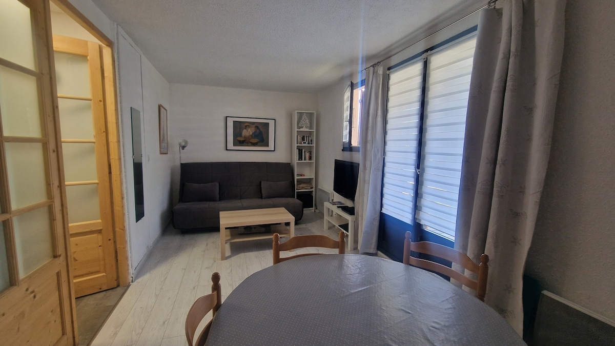 A living area features a dark sofa paired with a light wooden coffee table. A television is positioned on a shelf next to the window, which is fitted with light-colored blinds. A round dining table with wooden chairs is set in the foreground, enhancing the space's functionality.