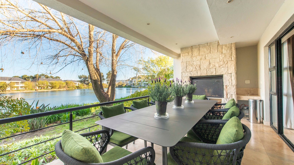 An outdoor patio area features a large dining table surrounded by comfortable green chairs. Potted plants are placed on the table, and a view of the nearby lake and trees can be seen through expansive sliding glass doors.