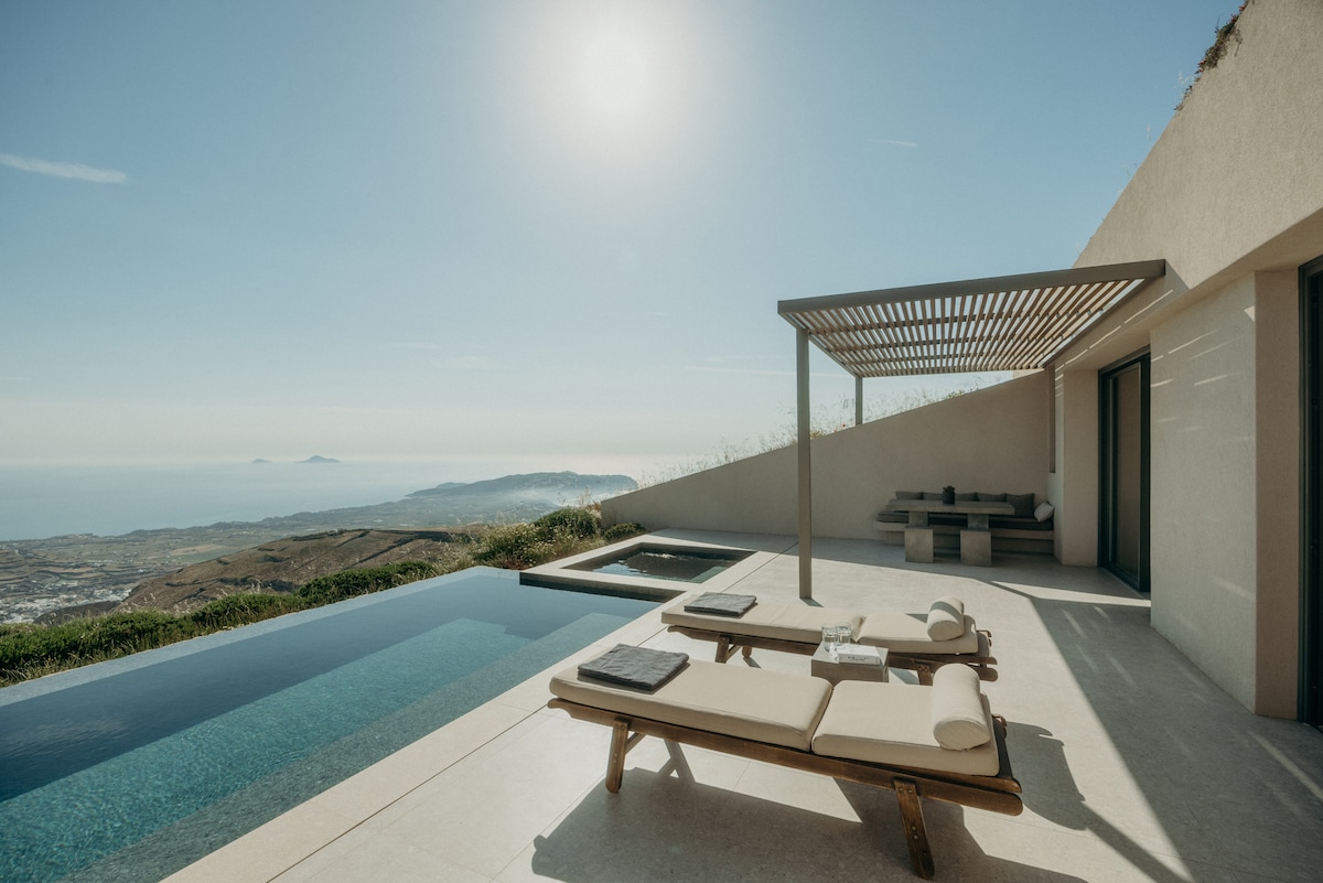A private terrace features a spacious infinity pool alongside a separate heated plunge pool. Comfortable lounge chairs are positioned by the pool, and a shaded dining area is visible under a modern pergola. Clear views of the sea and distant landscapes are offered.
