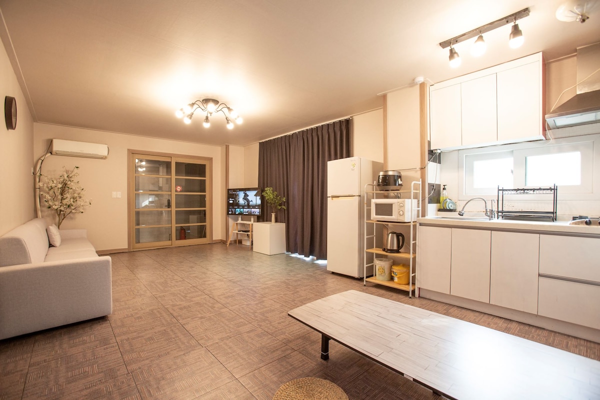 The spacious living area features a comfortable sofa facing a television and large glass doors that open to a private terrace. A modern kitchen, equipped with a refrigerator and cooking facilities, is visible, along with a dining table and decorative plants adding a touch of warmth.