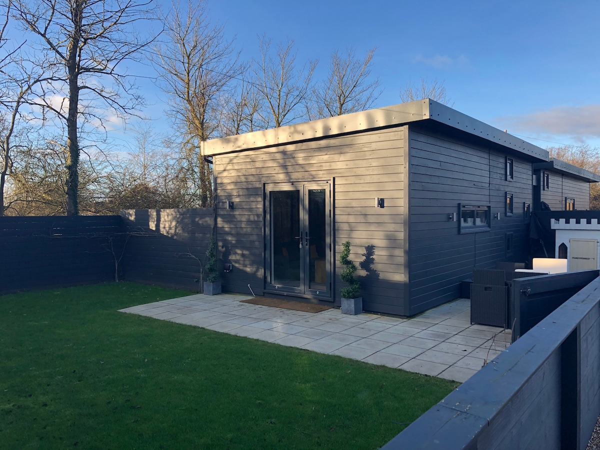 A modern detached studio is set amidst a private garden, featuring a stone-paved patio in front of large glass doors. The building's dark exterior contrasts with the lush green lawn and is surrounded by minimalist landscaping.