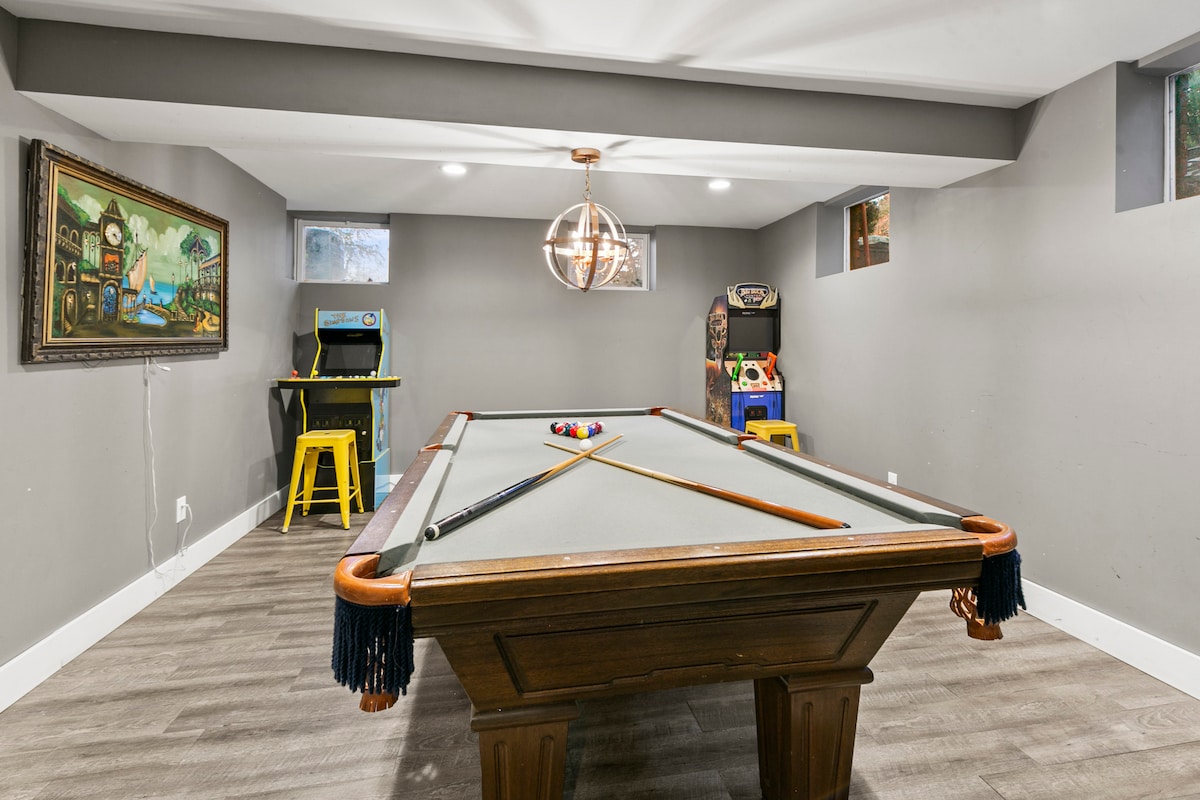A spacious game room features a billiard table at its center, surrounded by grey walls and modern lighting. An arcade game and a small table with yellow stools add vibrant accents to the space, while natural light filters in through small windows.