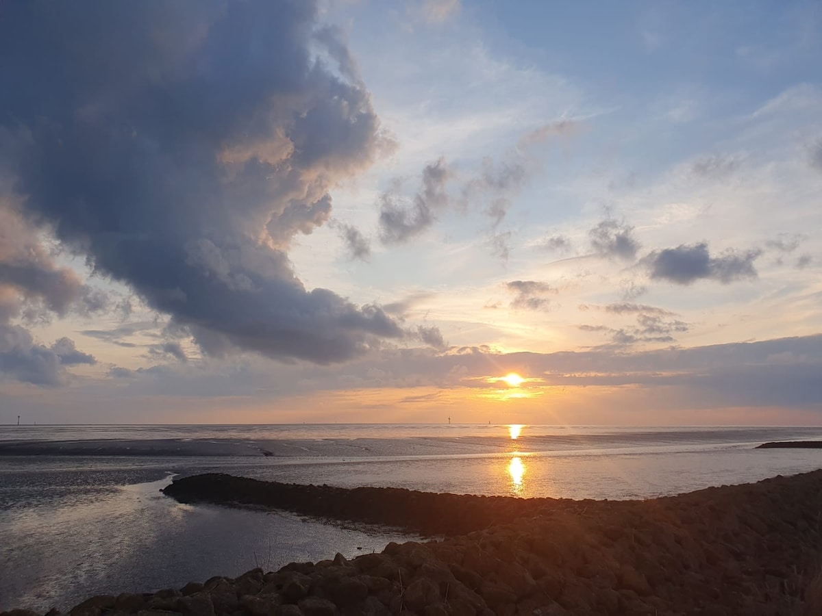 A serene sunset is captured over a tranquil waterway, with soft clouds scattered across the sky. The sun is gently setting on the horizon, illuminating the water. A rocky shoreline is visible in the foreground, providing a natural frame to the scene.