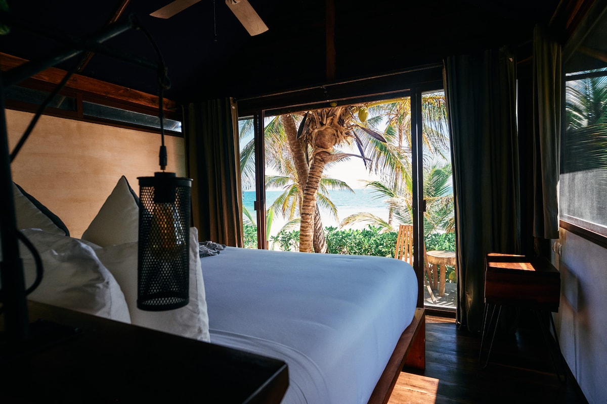 A spacious sleeping area features a large bed with soft linens, complemented by cozy pillows. Expansive glass doors provide a stunning view of palm trees and the ocean beyond, allowing natural light to flow into the room.