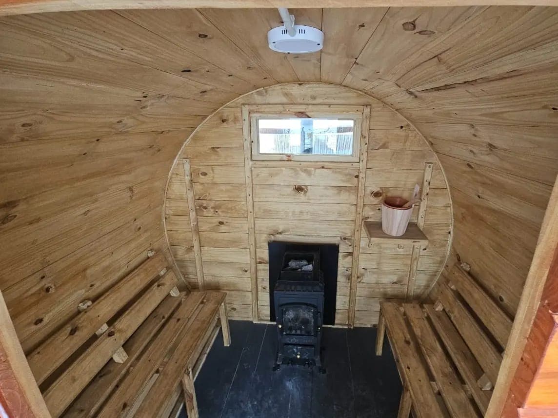 A cozy interior of a wooden structure is showcased, featuring two wooden benches and a compact wood-burning stove. Natural wood paneling creates a warm atmosphere, while a small window provides natural light. A shelf is mounted on the wall, adding functional space.